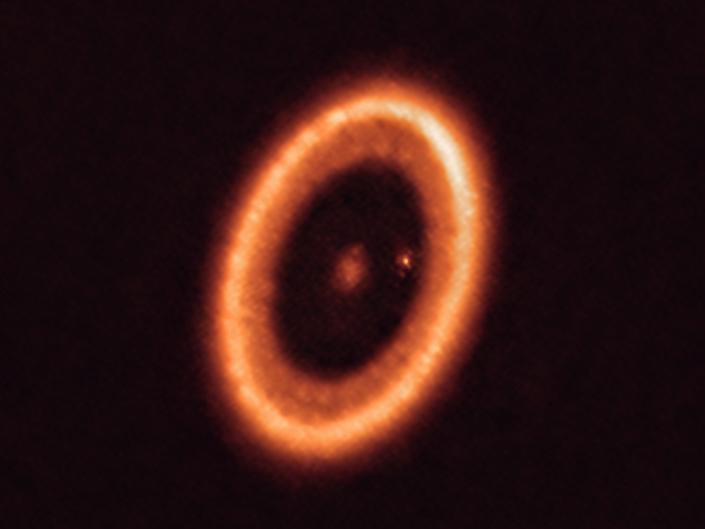 Gambar tersebut menunjukkan bintang oranye yang dikelilingi oleh piringan material oranye dengan planet bertitik kecil